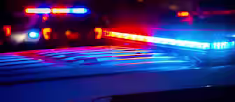 Close-up of a police car with a flashing red and blue light bar at night. Blurred lights from other emergency vehicles are visible in the background, casting a mix of blue and red hues across the scene. The atmosphere appears tense and urgent.