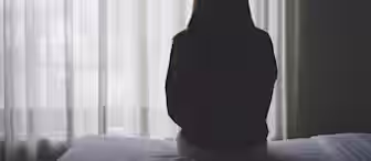 A person with long hair sits on the edge of a bed, facing away from the camera, looking out through a sheer curtain window. The room has soft lighting, creating a contemplative and serene atmosphere.