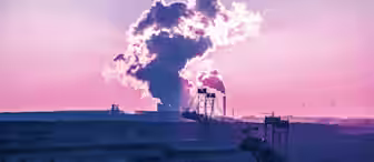 A dramatic scene of an industrial landscape at sunrise or sunset, with large excavators in a quarry in the foreground and massive plumes of smoke or steam rising from a distant facility against a purple-hued sky. Wind turbines are visible on the horizon.