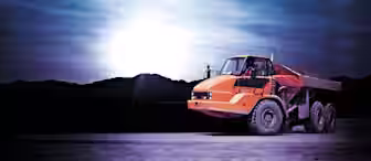 An orange dump truck on a dirt road at twilight. The truck's body casts shadows beneath it, while the setting sun creates a bright halo effect in the background, with dark mountains silhouetted against the sky.