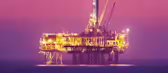 An offshore oil rig brightly lit against a serene dusk sky with a gradient from pink to orange. The rig stands alone in the middle of the calm ocean, with cranes and machinery visible. The water reflects the orange lights from the structure.