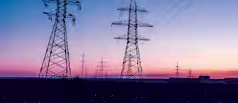 Several high-voltage electricity transmission towers and power lines stretch across a landscape at dusk. The sky is transitioning from pink to deep blue, enhancing the silhouette of the towers and wires. Buildings are faintly visible on the horizon.