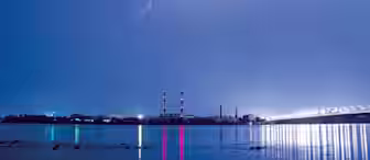 A nighttime scene shows lightning illuminating a cloudy sky over an industrial area. Two tall chimneys with red and white stripes stand prominently. Their reflections, along with numerous lights from the area, shimmer on the calm water below.