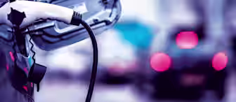A close-up of an electric vehicle being charged, with the charging cable plugged into the car's port. The background features out-of-focus cars driving on a city street, creating a blurred effect with red taillights.
