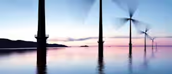 A serene ocean scene at dusk shows a row of large wind turbines with blades spinning against the colorful sky. The turbines are slightly blurred due to movement, while the calm water reflects the structures and the soft hues of the sunset.