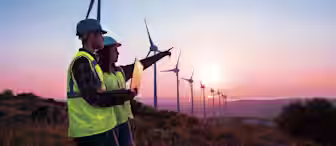 Two engineers wearing safety helmets and reflective vests stand in front of several wind turbines at sunset. One holds a laptop while the other points towards the wind farm. The sky is a blend of pink, orange, and blue hues.