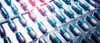 Close-up of several blister packs filled with blue and white capsules. The capsules are arranged in a grid-like pattern, and the lighting highlights some of the rows, giving a metallic sheen to the packaging.