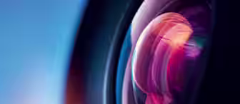 Close-up shot of a camera lens reflecting colorful light. The lens exhibits a spectrum of colors including pink, purple, and blue, with a blurred, gradient blue background that transitions from dark to light. The image emphasizes the intricate details of the lens.