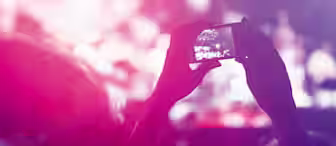 A person holding up a smartphone to capture a concert or event. The background is blurred with vibrant pink, purple, and white lights, giving a festive and energetic atmosphere. The image focuses on the phone screen where a part of the stage is visible.
