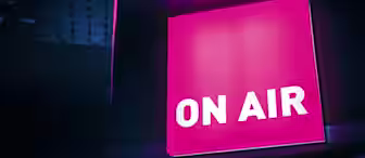 Illuminated rectangular sign with the words "ON AIR" in bold white letters against a vibrant pink background. The sign is fixed to a dark ceiling, creating a high-contrast look with the neon glow casting light into the surrounding space.