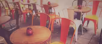 Outdoor café with round wooden tables and vibrant orange and white metal chairs. Each table has a red decorative candle holder. The setting is peaceful, with a cobblestone ground and a soft, warm light filtering through.