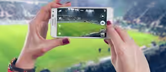 A person with red-painted nails is holding a smartphone, capturing a photo of a soccer field in a large stadium. The stands are filled with spectators, and teams are playing on the brightly lit field.