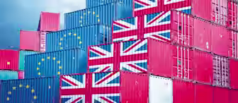 A stack of shipping containers decorated with the flags of the United Kingdom and the European Union against a cloudy blue sky. The containers are arranged in a stepped formation, with some bearing the UK flag and others the EU flag.