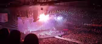 A vibrant concert scene with a large crowd. The stage is bathed in colorful lights with multiple spotlights focusing on the performers. The audience is cheering and waving their hands in the air. Confetti is falling from above, enhancing the festive atmosphere.