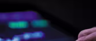 A hand is interacting with a tablet displaying a financial graph with fluctuating lines and numbers. The screen shows a dark background with bright purple, green, and white elements, depicting stock or market analytics.