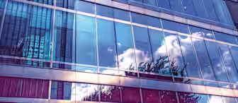 A modern skyscraper with a glass facade reflects the sky, clouds, and neighboring buildings. The image is taken from a low angle, emphasizing the height and sleek design of the structure. The blue tones create a serene, contemporary atmosphere.
