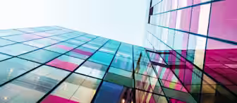 A low-angle view of modern glass buildings reflecting colorful hues of pink, red, blue, and yellow under a clear sky. The structures create a geometric pattern with their intersecting lines and vibrant panels.