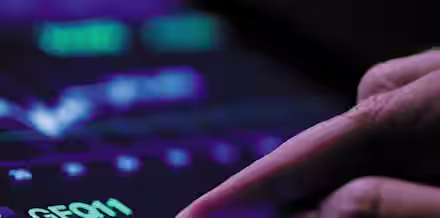 A hand is interacting with a tablet displaying a financial graph with fluctuating lines and numbers. The screen shows a dark background with bright purple, green, and white elements, depicting stock or market analytics.