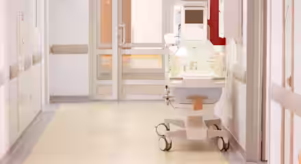 A hospital hallway with a neonatal incubator on wheels positioned near a closed door, under bright overhead lighting, and surrounded by white walls and medical equipment.