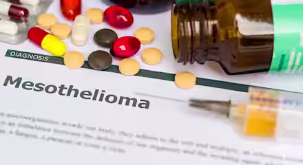 A document titled "Mesothelioma" is surrounded by various medications, including pills and capsules, and an open pill bottle. A syringe rests on the paper.