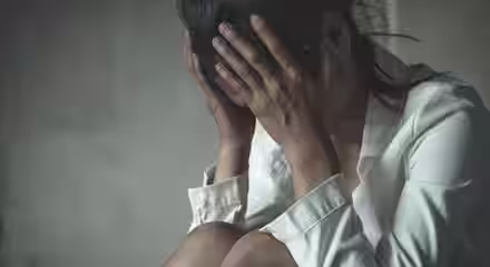 A person with dark hair, wearing a white shirt, is seated with their head in their hands, appearing distressed. The background is blurry, and the overall color tone of the image is muted, conveying a sense of sadness or contemplation.