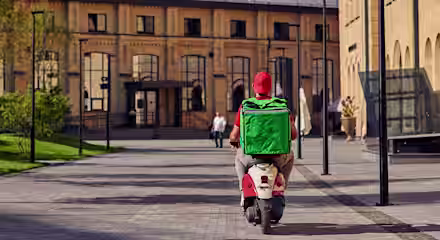 A food delivery person wearing a red cap and shirt rides a scooter with a large green insulated bag on their back through a modern urban courtyard with brick buildings.