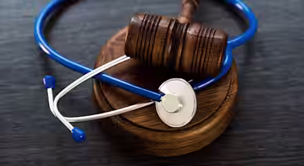 A wooden judge’s gavel resting on a circular sound block with a blue stethoscope draped around it, symbolizing the intersection of law and healthcare.