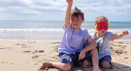 Two young children sit on a sandy beach by the ocean under a blue sky. One child has short brown hair, wearing a blue shirt and denim shorts, and raises an arm in the air. The other child wears a superhero mask and also raises an arm, mimicking flying.