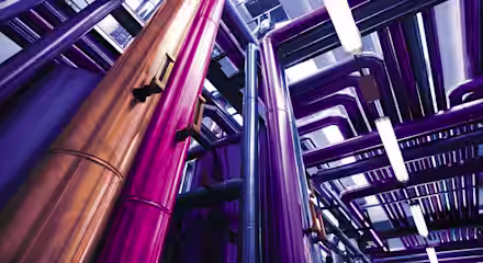 A vibrant and colorful industrial scene with tall metallic pipes in shades of purple, pink, and copper running in various directions under bright lighting. The pipes are part of a complex system, creating a visually striking and futuristic environment.