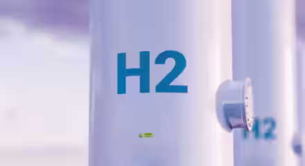 Close-up of a white cylindrical tank with "H2" written in blue, indicating hydrogen storage. The tank has a small valve or connector on the side. The background is slightly blurred with a light pinkish hue sky.