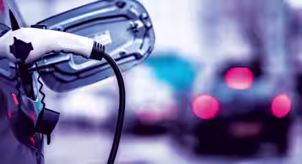 A close-up of an electric vehicle being charged, with the charging cable plugged into the car's port. The background features out-of-focus cars driving on a city street, creating a blurred effect with red taillights.