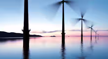 A serene ocean scene at dusk shows a row of large wind turbines with blades spinning against the colorful sky. The turbines are slightly blurred due to movement, while the calm water reflects the structures and the soft hues of the sunset.
