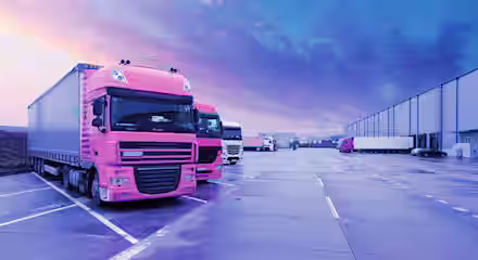 A row of colorful semi-trucks, primarily pink, is parked on a vast, wet pavement near a distribution warehouse. The sky above is a mix of purple and blue hues, suggesting dusk or dawn, with a serene ambiance reflecting off the ground.