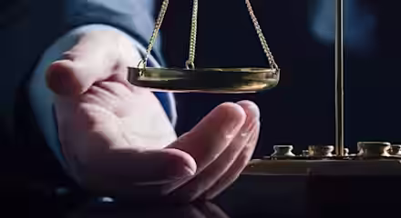 A person’s hand reaches toward a brass balance scale on a dark reflective surface, symbolizing justice and fairness, with a dark, blurred background.