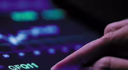 A hand is interacting with a tablet displaying a financial graph with fluctuating lines and numbers. The screen shows a dark background with bright purple, green, and white elements, depicting stock or market analytics.