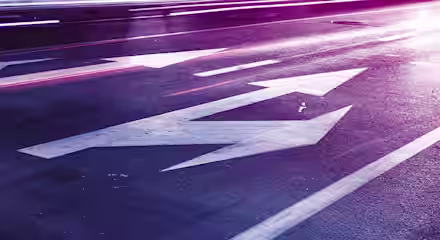 A nighttime street scene with multiple painted white arrows on the pavement, pointing in different directions. The street and arrows are illuminated by vibrant, colorful lights, creating a surreal, almost neon effect. Blurred light trails from vehicles can also be seen.