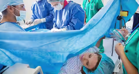 A medical team in surgical scrubs performs a procedure in an operating room. A patient lies on the operating table with a blue surgical drape placed over her lower body. Nearby, a nurse holds an anesthesia mask near the patient’s face.