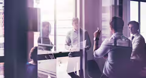 Group of five business people in a modern office with large windows. They are having a discussion around a table, with one person standing and holding papers, another gesturing with a pen, and the rest seated. The room is filled with natural light.