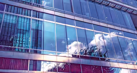 A modern skyscraper with a glass facade reflects the sky, clouds, and neighboring buildings. The image is taken from a low angle, emphasizing the height and sleek design of the structure. The blue tones create a serene, contemporary atmosphere.