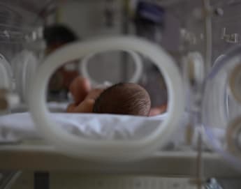 A newborn baby lies on its stomach inside a hospital incubator. The focus is on the baby's head, with soft lighting and a blurred background. The incubator's round openings surround the infant, providing a glimpse into the care setting.