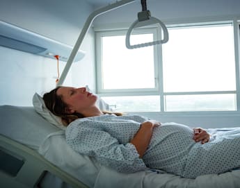 A woman in a hospital gown lies on a bed in a brightly lit hospital room, holding her pregnant belly. She appears to be in a restful state, looking up towards the ceiling. A support handle hangs above her from an overhead bar.