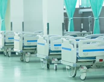 A row of empty hospital beds with white sheets and blue accents are lined up against a wall in a brightly lit room. Green privacy curtains are partially drawn between each bed, and the floor is clean and shiny. The atmosphere appears sterile and well-maintained.