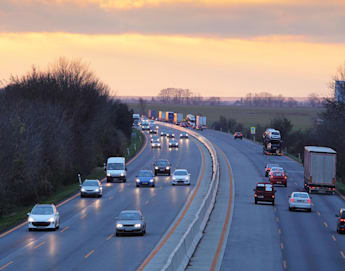 A highway at sunset with a mix of cars and trucks traveling in both directions. The left side of the road is busier with several vehicles, while the right side has fewer vehicles. Trees border the highway on the left, and open fields are visible on the right.