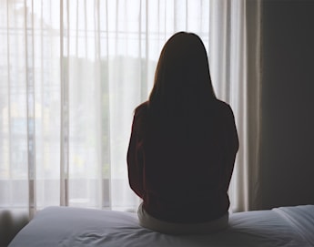 A person with long hair sits on the edge of a bed, facing away from the camera, looking out through a sheer curtain window. The room has soft lighting, creating a contemplative and serene atmosphere.