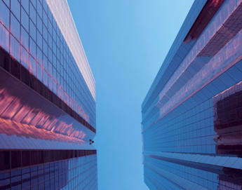 A perspective view looking up between two modern glass skyscrapers against a clear blue sky. The reflections on the glass panes give the buildings a slightly pinkish tint. The symmetrical structures create a dramatic, towering geometric effect.