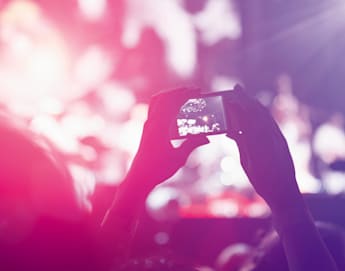 A person holding up a smartphone to capture a concert or event. The background is blurred with vibrant pink, purple, and white lights, giving a festive and energetic atmosphere. The image focuses on the phone screen where a part of the stage is visible.