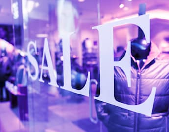 A store window with large white letters spelling "SALE" in a brightly lit retail environment. Inside, blurred mannequins display winter jackets. The scene has a purple-tinted lighting effect.