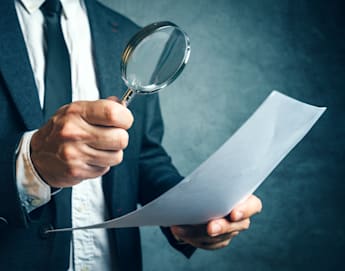 A person wearing a suit and tie is holding a magnifying glass in their right hand and examining a sheet of paper in their left hand. The background is blurred and has a dark, textured appearance.