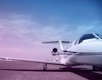 A sleek private jet is parked on an open tarmac under a clear sky with a gradient of purple and blue hues. The jet's wings extend outward, and the view focuses on the front portion and one of the engines. The landscape is open and expansive.