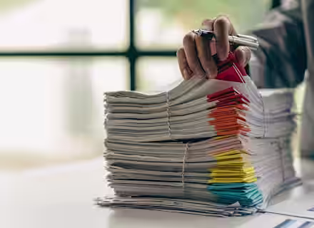 A person organizing a tall stack of paper documents with colorful tabs on a table. Their hand, holding a pen, is adjusting the papers. A large window in the background provides natural light.